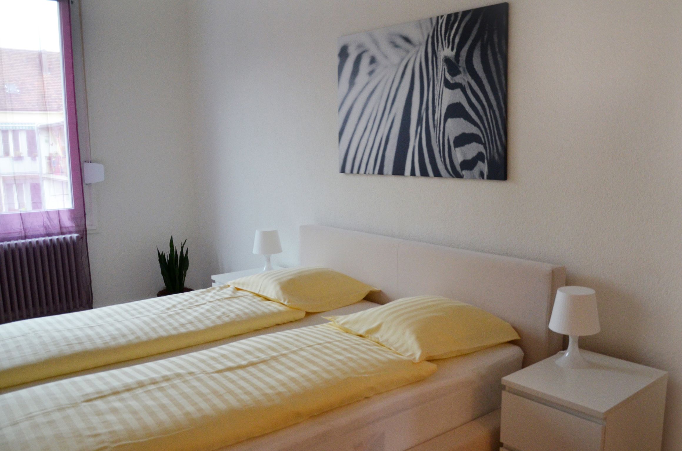 A hotel room with a bed, two bedside lamps, a zebra print artwork, a potted plant, and a window with blinds.