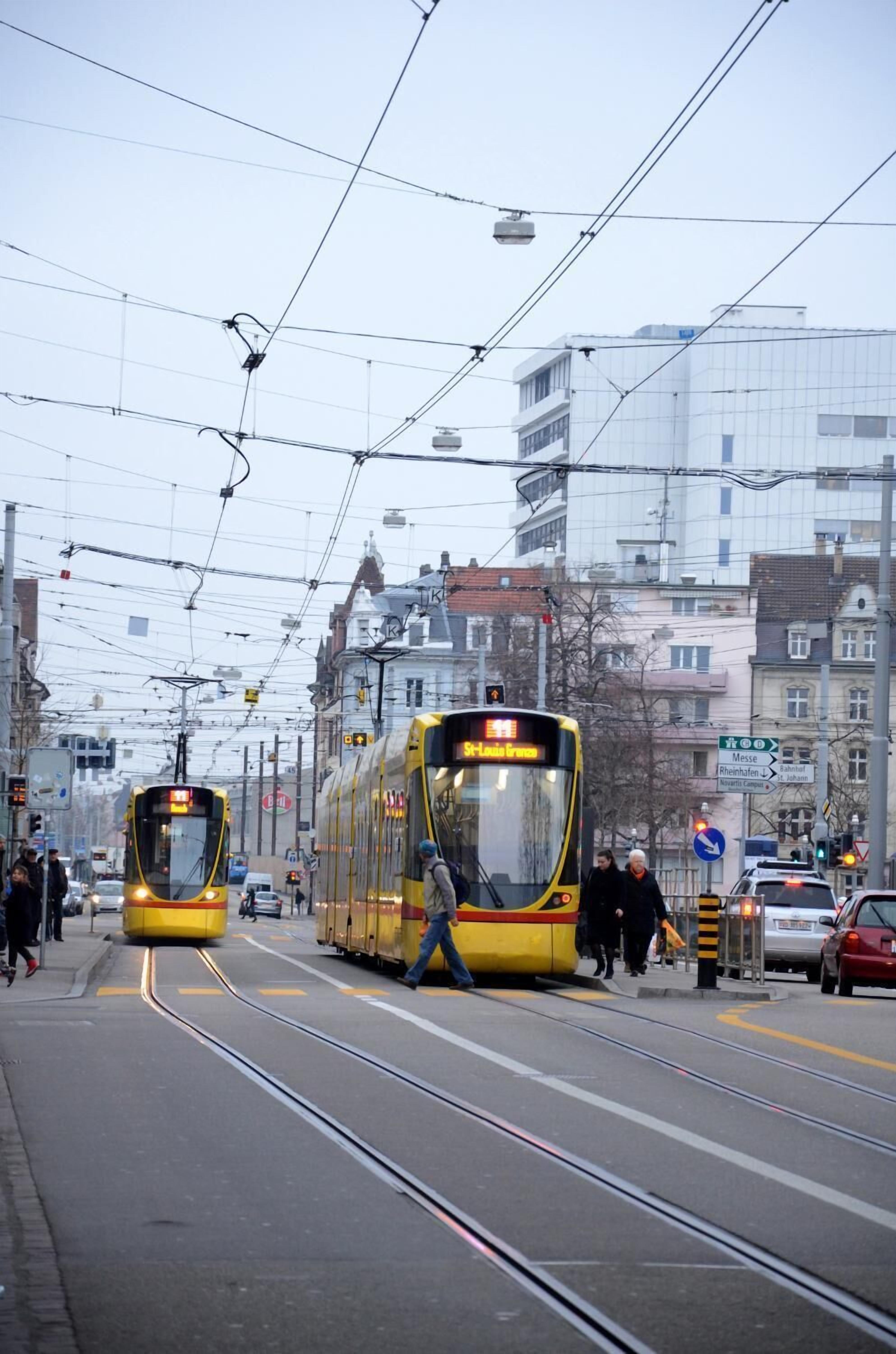 A tram station with two trams, overhead wires, and buildings in the background.