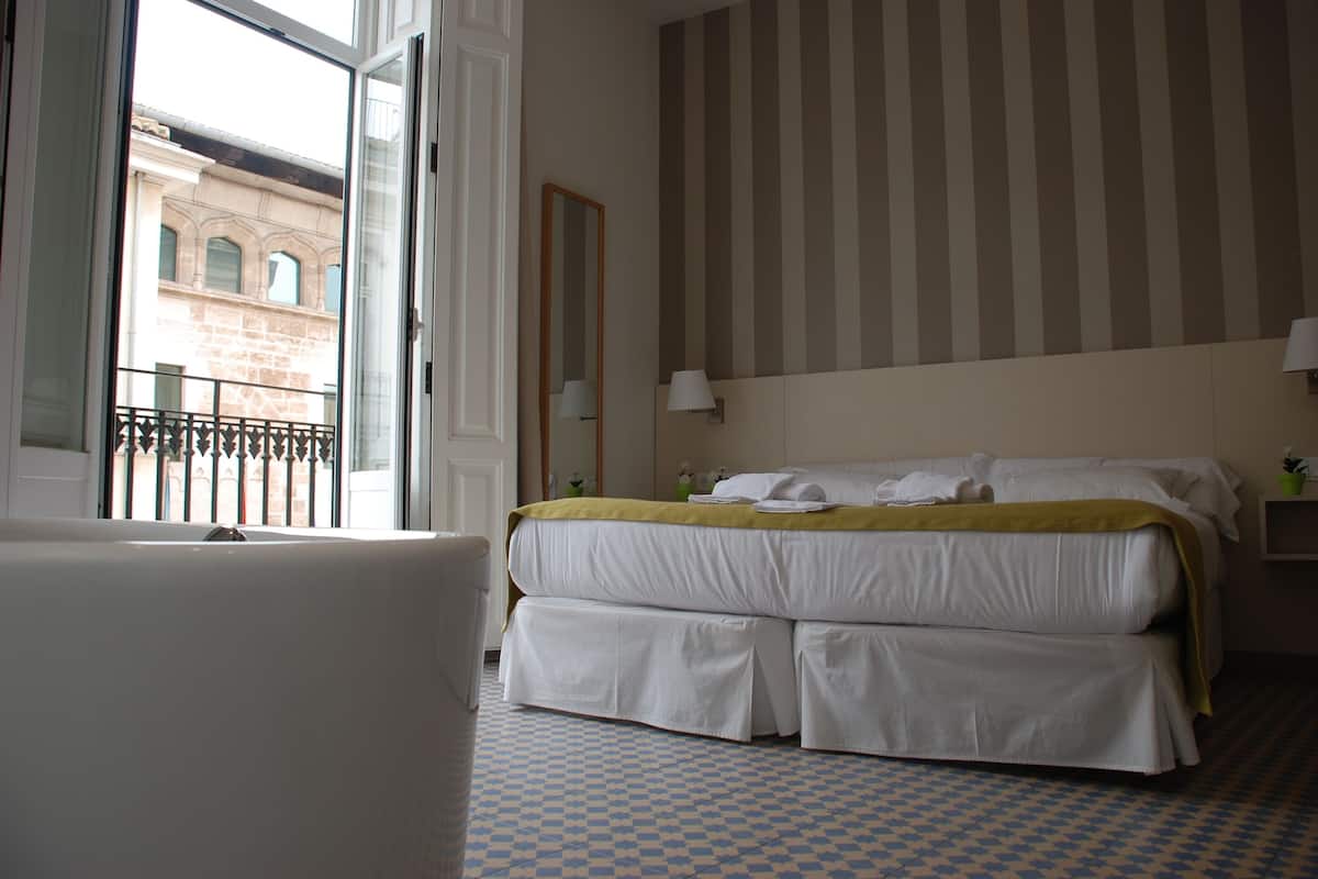 A hotel room with a large bed, a window with a view of a building, a mirror, and a checkered floor.