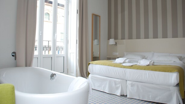 Quarto de hotel com uma banheira grande, uma cama de casal com roupa de cama branca, parede listrada e varanda com vista para edifícios.