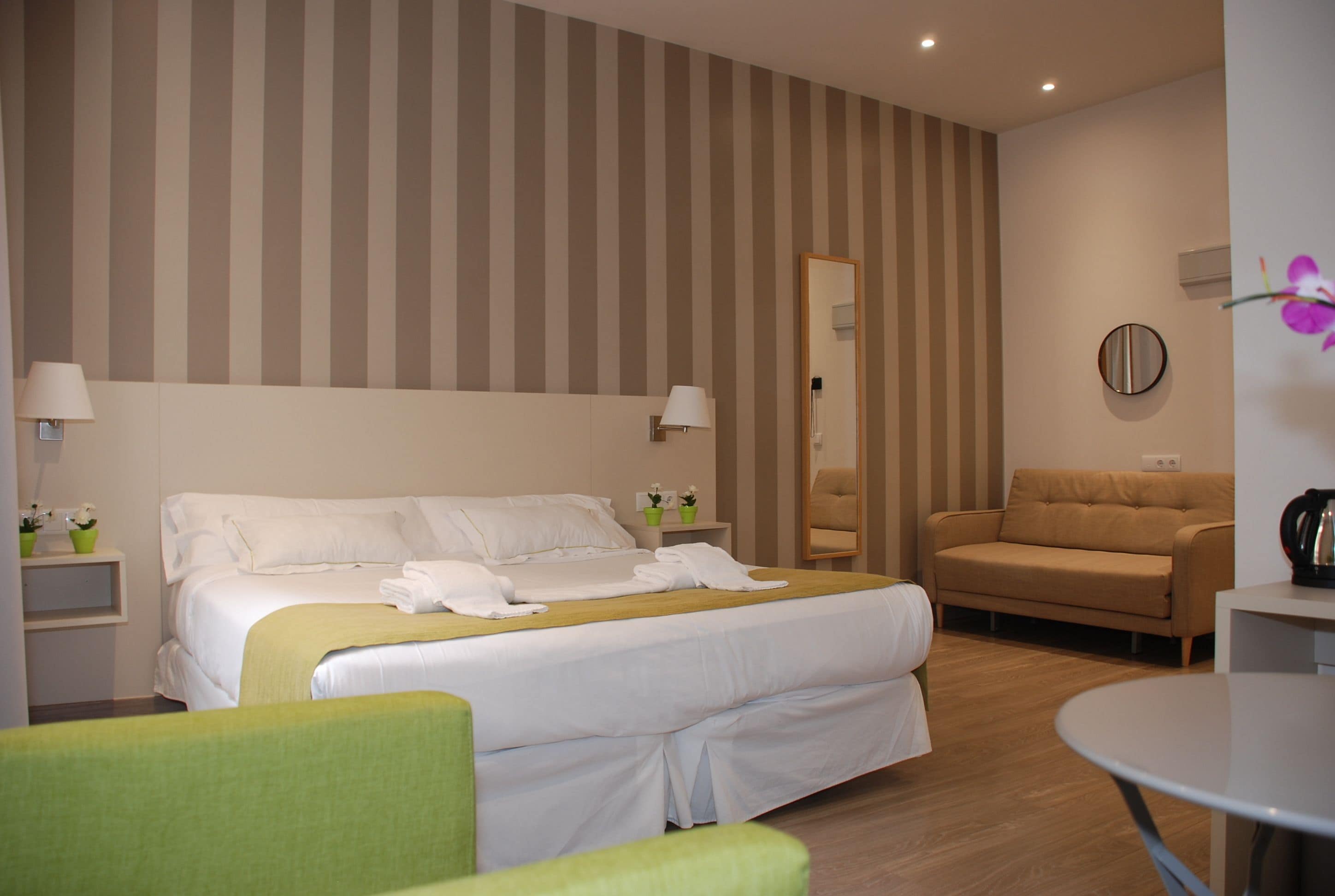 A hotel room with a bed, a green armchair, a sofa, a round table, and a mirror on the wall.