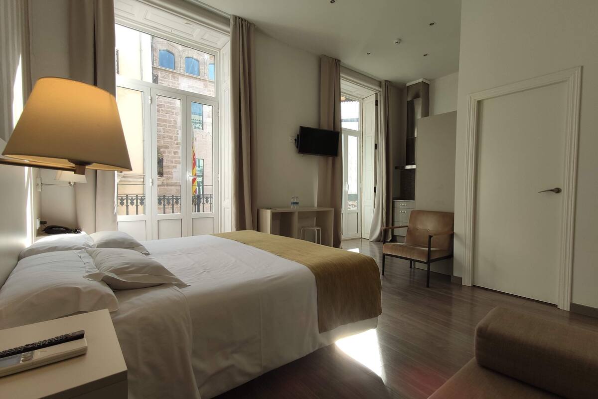 A hotel room with a bed, a desk, a chair, a TV, and a balcony with a view of buildings.