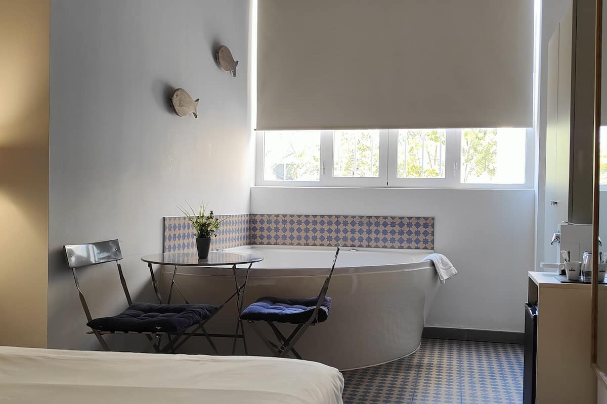 A hotel room with a bed, a small table with chairs, a window with blinds, a bathtub, and a wall-mounted lamp.