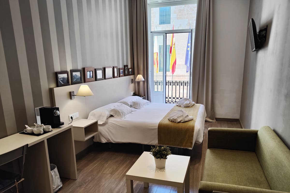 A hotel room with a bed, bedside table, desk, and a small table with a plant.