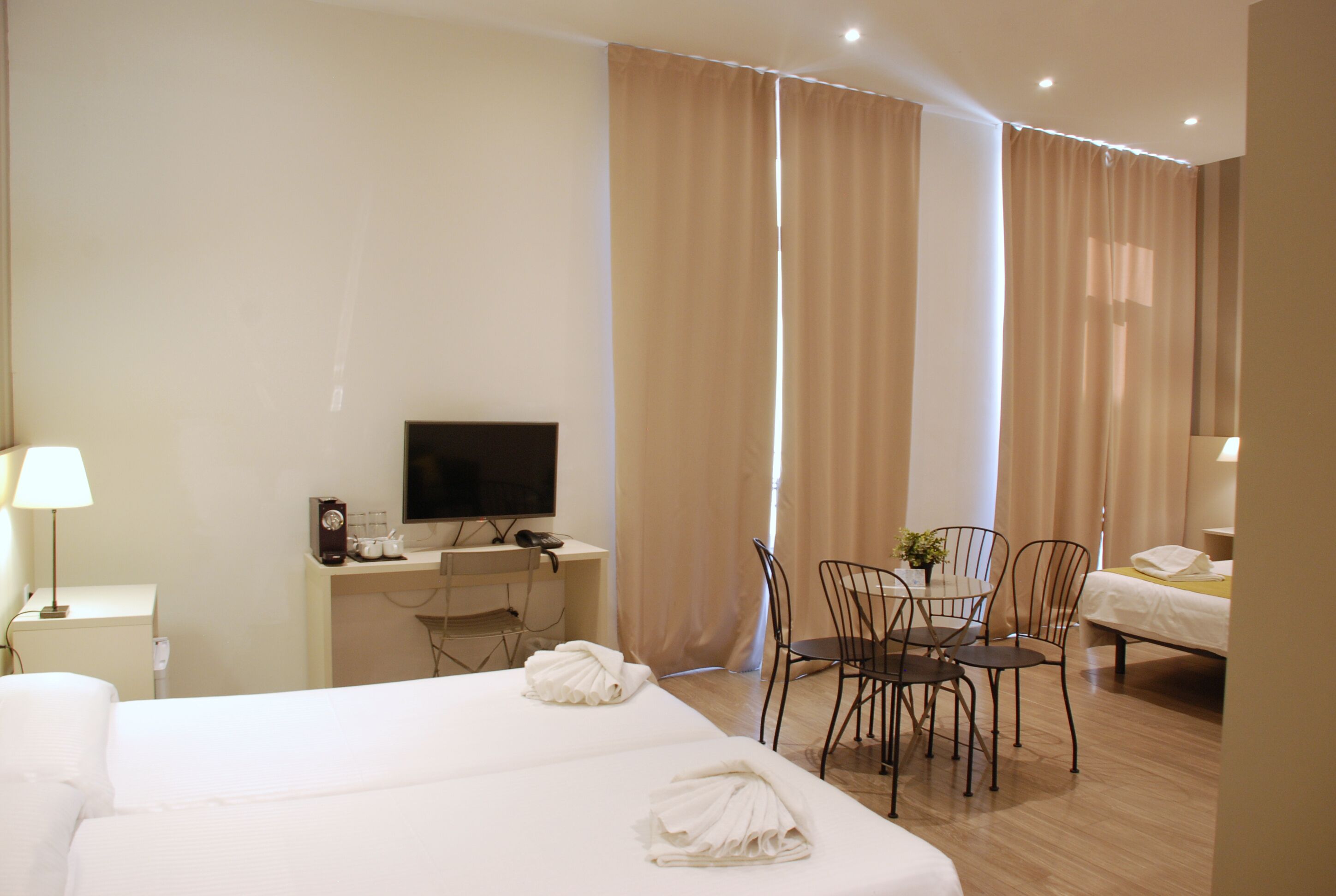 Habitación de hotel con dos camas, un escritorio con computadora, televisor, mesa de comedor con sillas y ventana con cortinas.