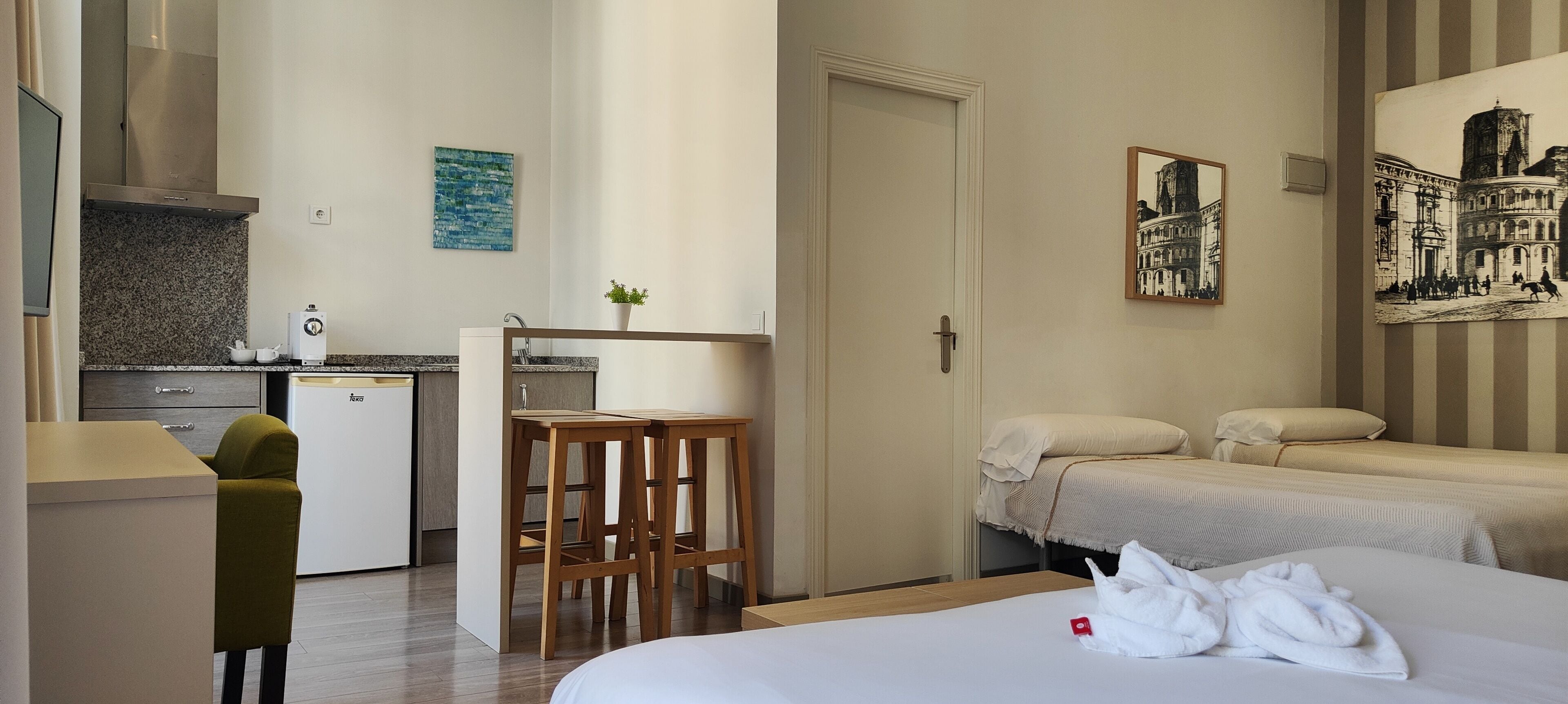 Habitación de hotel con kitchenette, una cama con dos almohadas, una mesita con taburetes y obras de arte en las paredes.