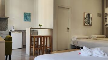 Habitación de hotel con kitchenette, una cama con dos almohadas, una mesita con taburetes y obras de arte en las paredes.