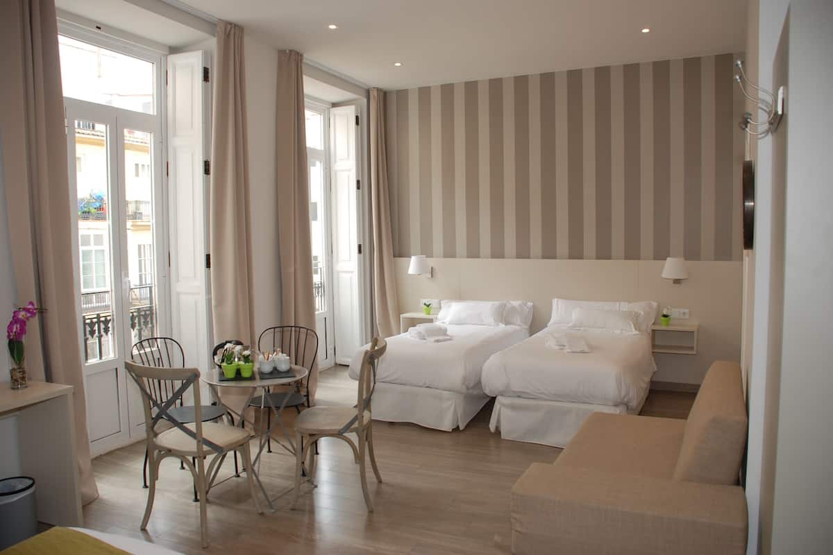 A hotel room with two beds, a sofa, a dining table with chairs, and a balcony with curtains.