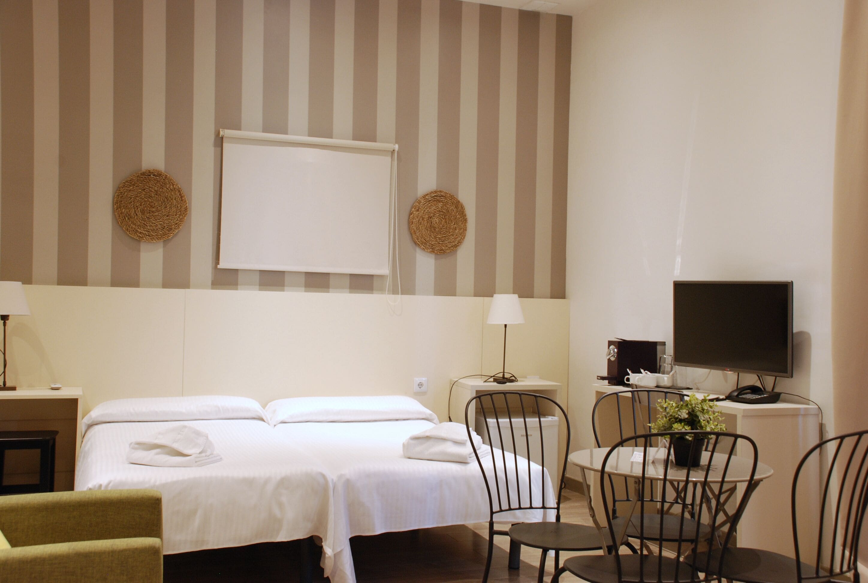 A hotel room with two beds, a desk, a chair, a small table, a TV, and two wall-mounted lamps.