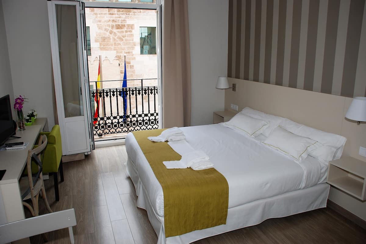 A hotel room with a bed, a desk with a chair, a TV, and a balcony with a view of the city.