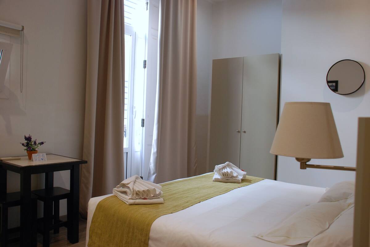 A hotel room with a bed, a desk, a chair, a lamp, a wardrobe, and a mirror.