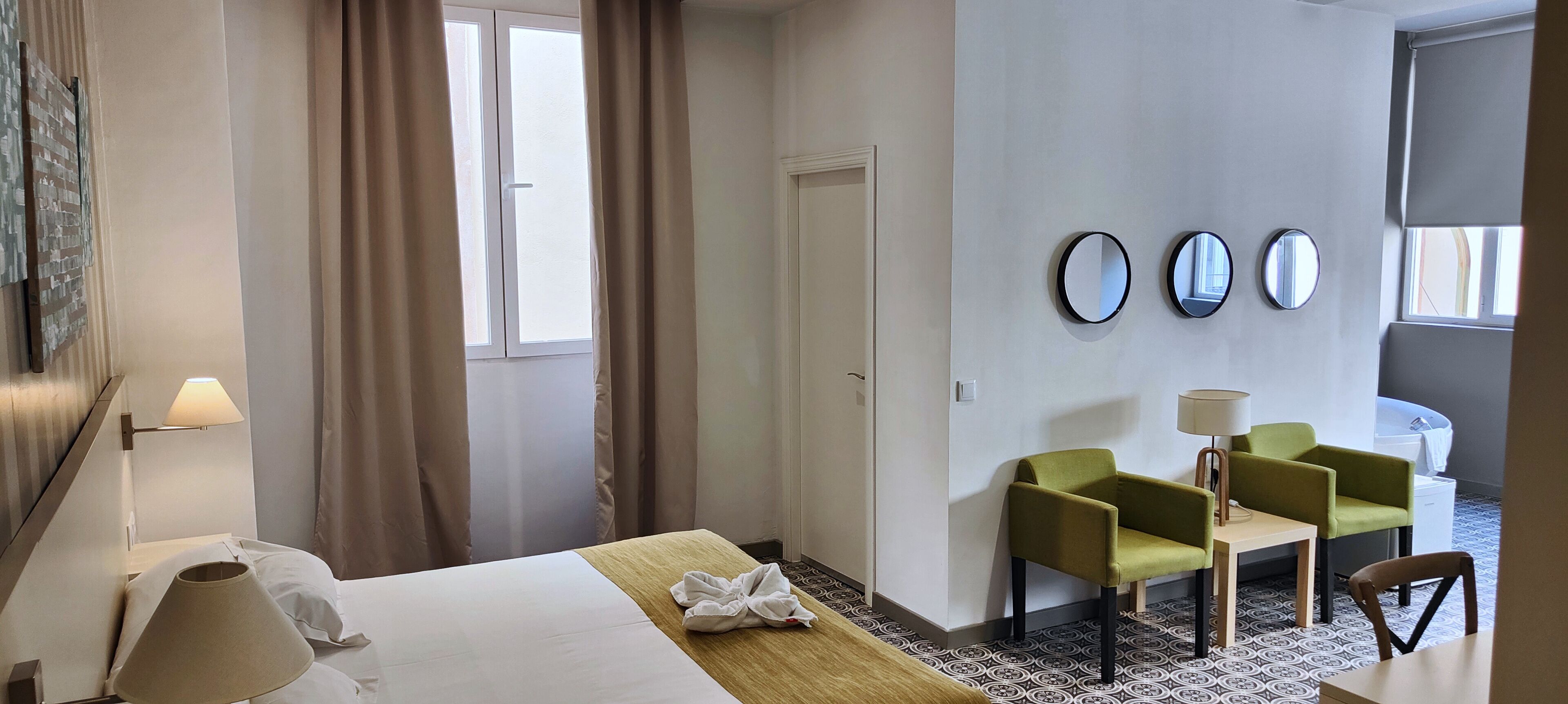 A hotel room with a bed, a desk, two chairs, a lamp, and a window with curtains.