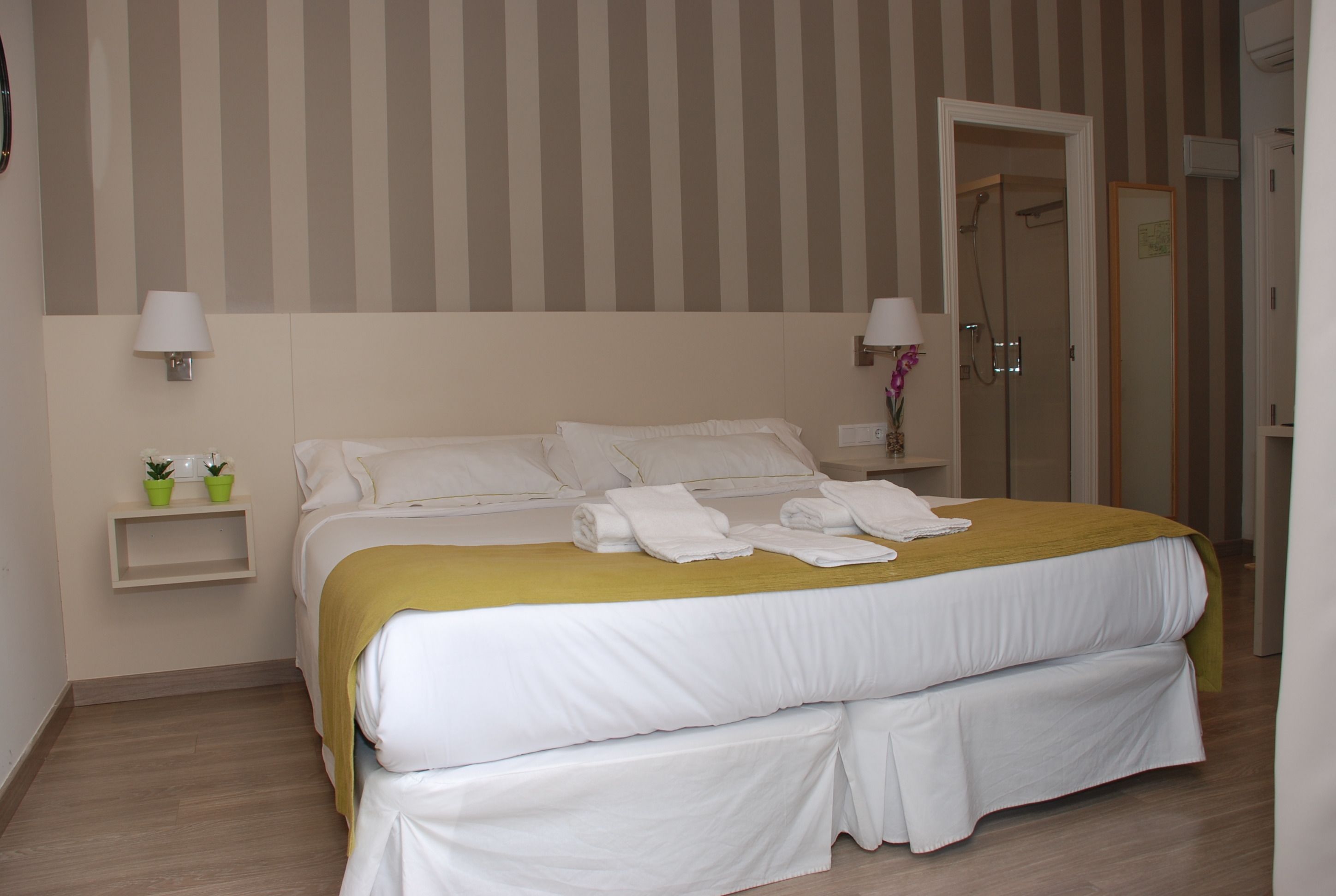 Cama doble con sábanas blancas, un plaid color mostaza y toallas dobladas con cuidado. Hay una mesita de noche con una lámpara y dos plantas en macetas. La habitación tiene un diseño de papel tapiz a rayas y suelo de madera.