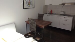 A compact hotel room with a bed, a small dining table with a chair, a kitchenette with a stove, and a framed picture on the wall. - rent-a-home Schweizergasse (Basel)