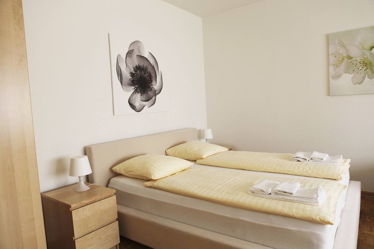 A double bed with two pillows, a bedside table with a lamp, and artwork on the wall.