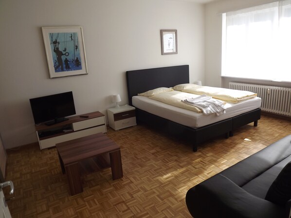 A bedroom with a bed, a TV, a bedside table, a lamp, and a framed picture on the wall. - rent-a-home Schweizergasse (Basel)