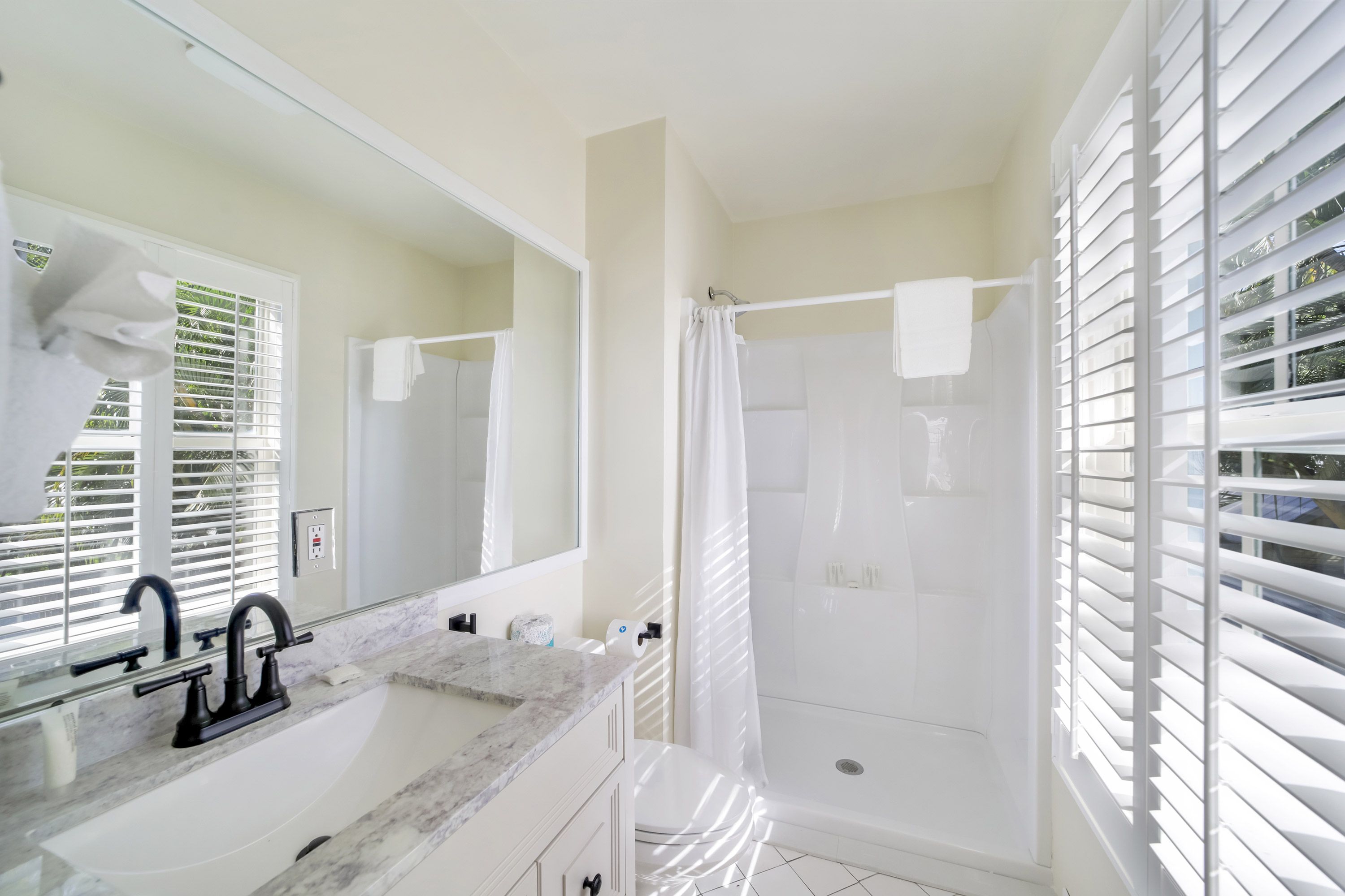 Un baño con lavamanos de mármol, grifo negro, ducha con cortina blanca y ventana con persianas.