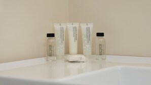 Un baño con un lavamanos blanco, tres botellas de loción corporal y una toalla blanca.