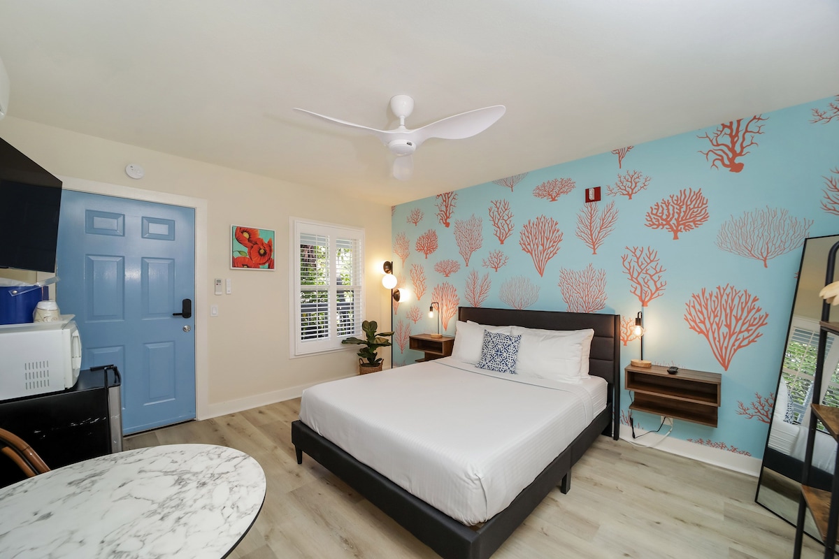 Habitación de hotel con cama, escritorio, silla, ventilador de techo y un muro con motivos de árbol de coral.