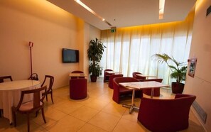 A conference room with a round table, red chairs, a TV, potted plants, and a large window with curtains.
