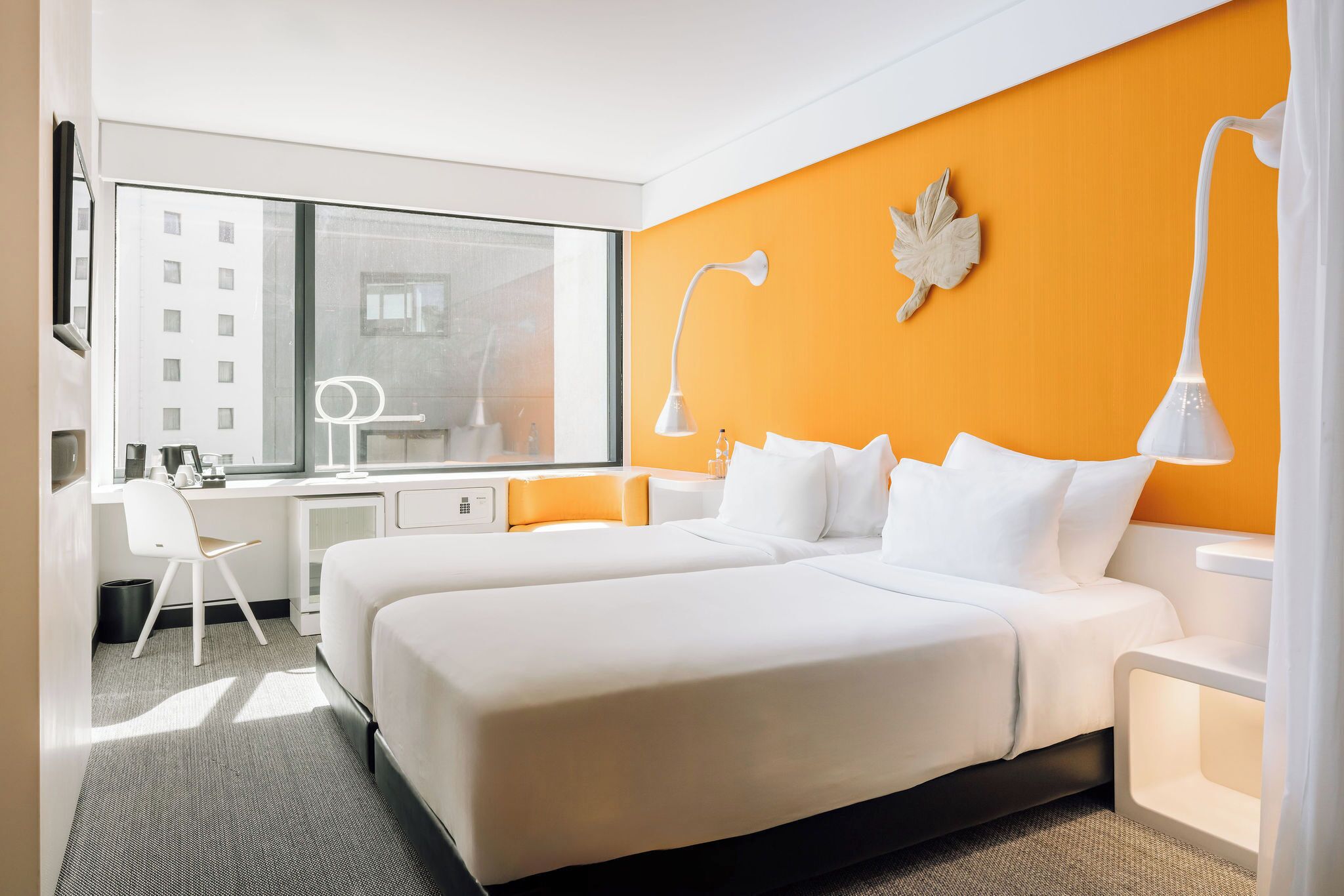 A hotel room with two beds, a desk, a chair, a large window, and a decorative leaf on the wall.