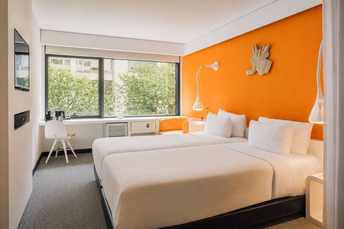 Habitación de hotel con un ventanal grande, un escritorio, dos camas y una pared de acento naranja.