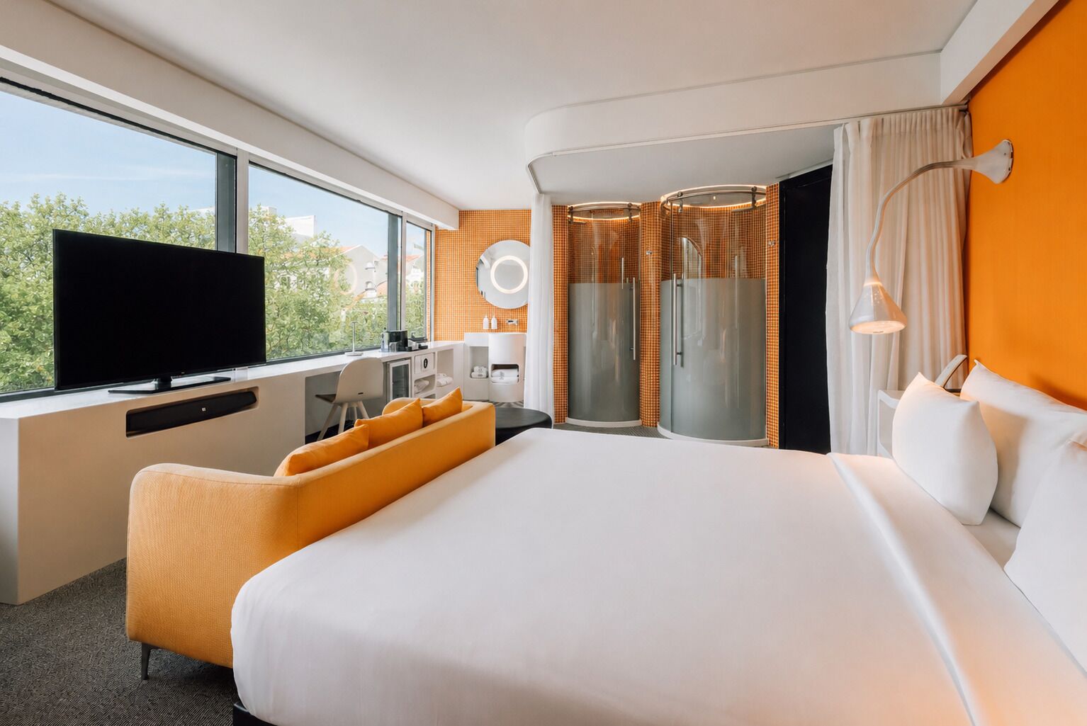 A modern hotel room with a large bed, a desk with a flat-screen TV, a seating area with orange cushions, a shower area, and a view of greenery through the window.