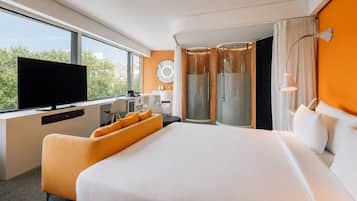 Ein modernes Hotelzimmer mit einem großen Bett, einem Schreibtisch mit Flachbildfernseher, einer Sitzecke mit orangefarbenen Kissen, einer Dusche und Blick ins Grüne durch das Fenster.
