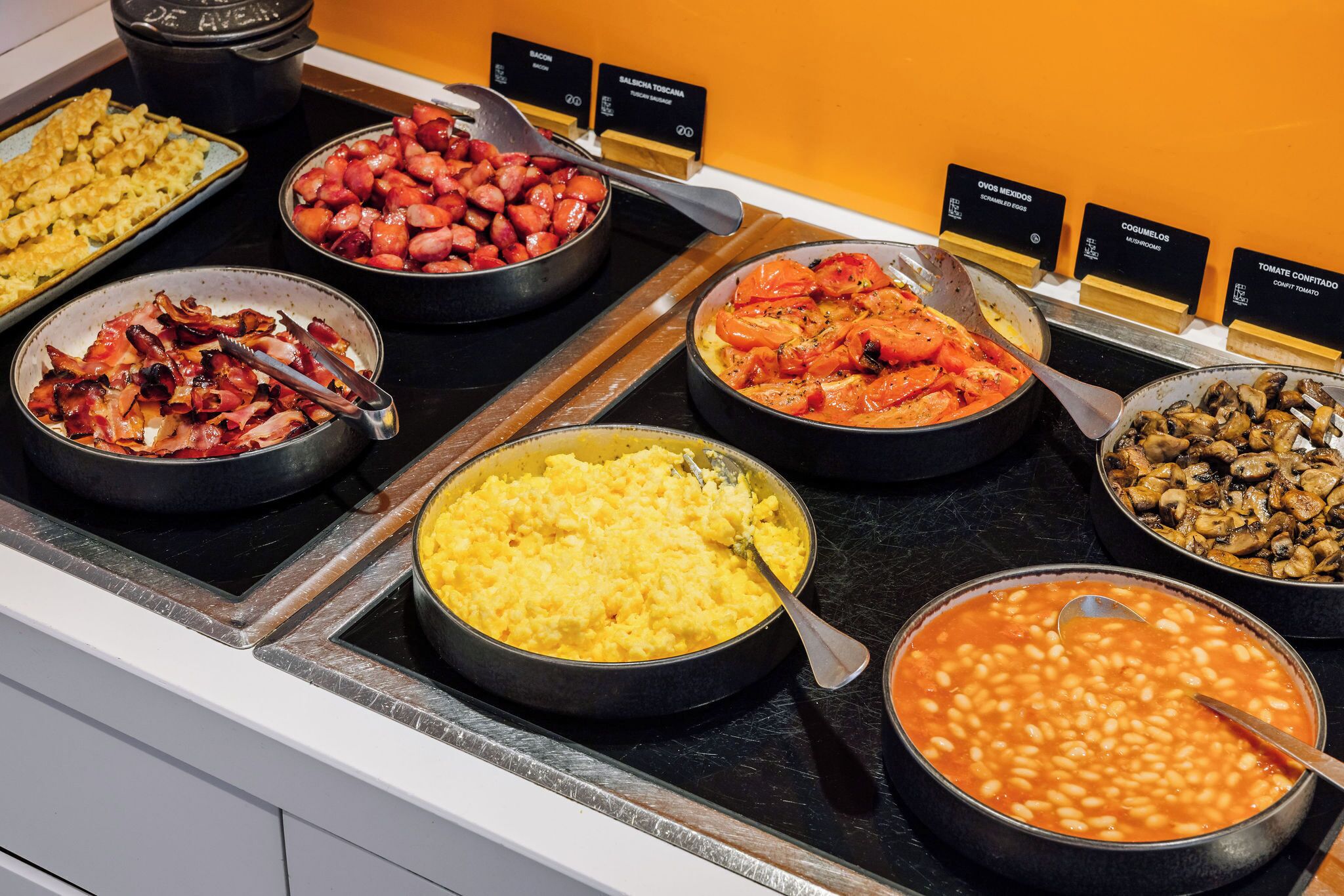 Un buffet proposant divers plats, notamment des œufs brouillés, du bacon, des saucisses, des tomates rôties, des champignons et des haricots.