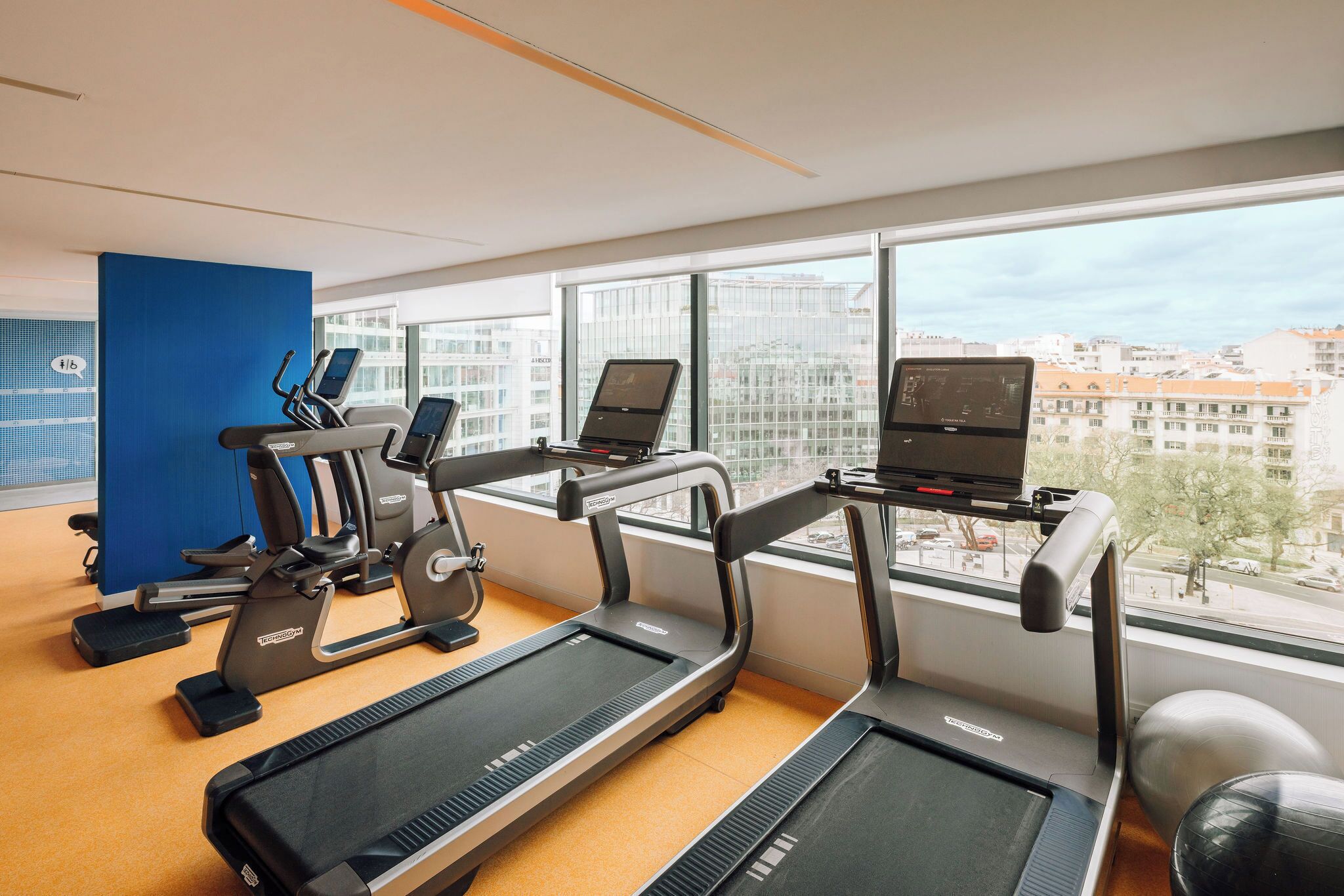 Un gimnasio con cintas de correr, una bicicleta estática y una máquina de remo, con amplias ventanas que ofrecen vista a la ciudad.