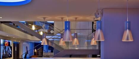 A modern hotel lobby with a spacious seating area, a staircase, and a large digital display wall.