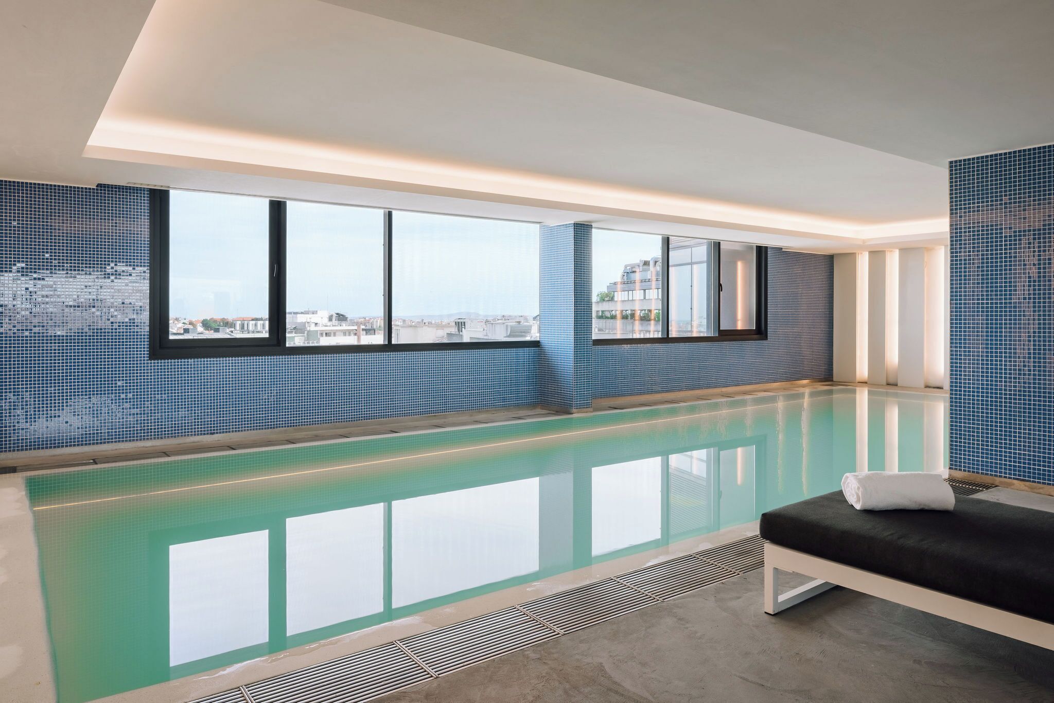 Una piscina interior moderna con vistas a la ciudad, un área de descanso con un banco y una pared de azulejos.