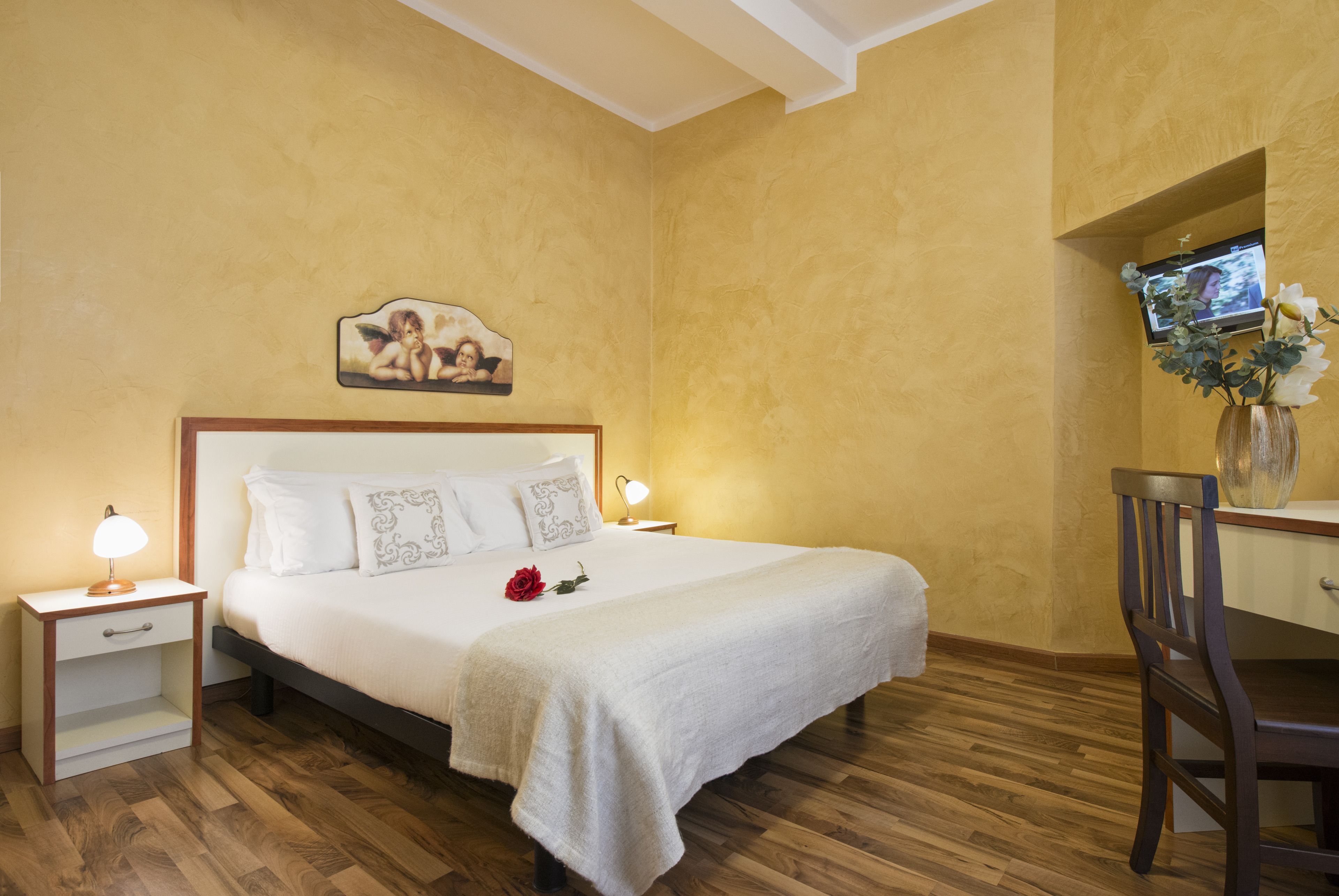 Uma cama bem arrumada com colcha e travesseiros brancos, uma mesa de cabeceira com abajur, piso de madeira e televisão fixada na parede.