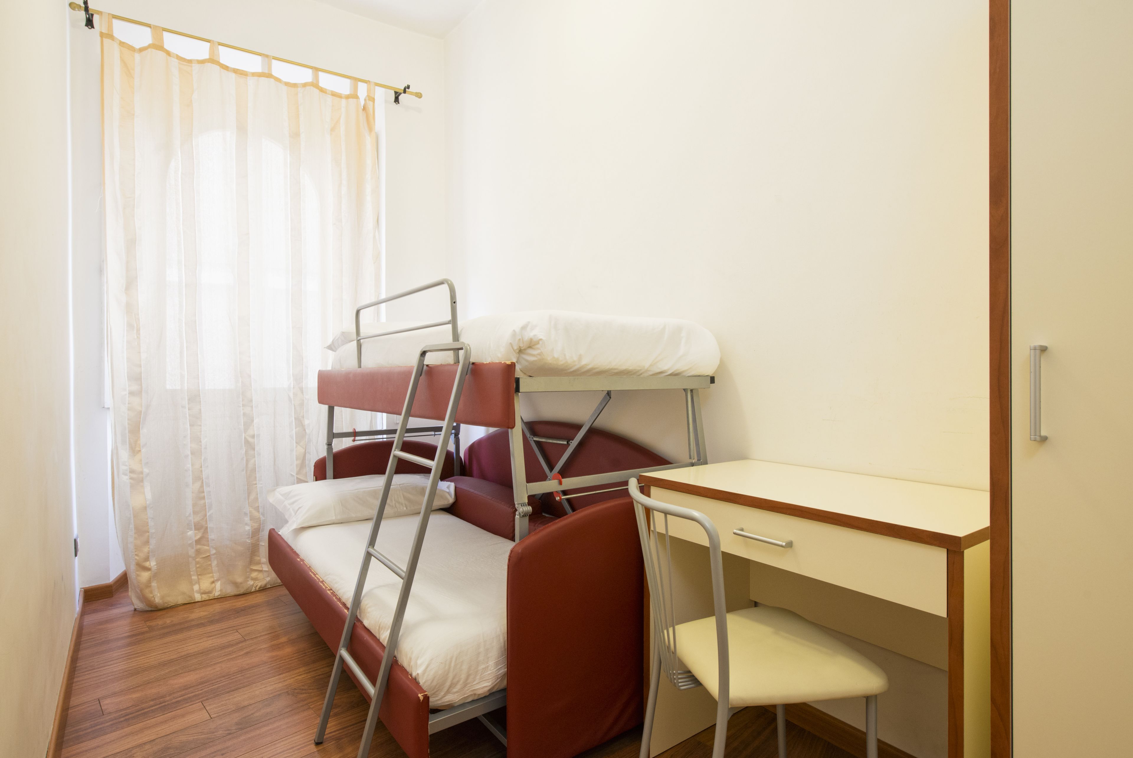 A bunk bed with a desk and chair, a window with sheer curtains, and a wooden floor.