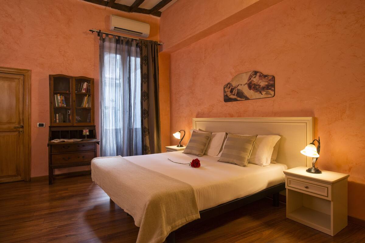 A bedroom with a large bed, bedside lamps, a nightstand, a wooden dresser, and a bookshelf.