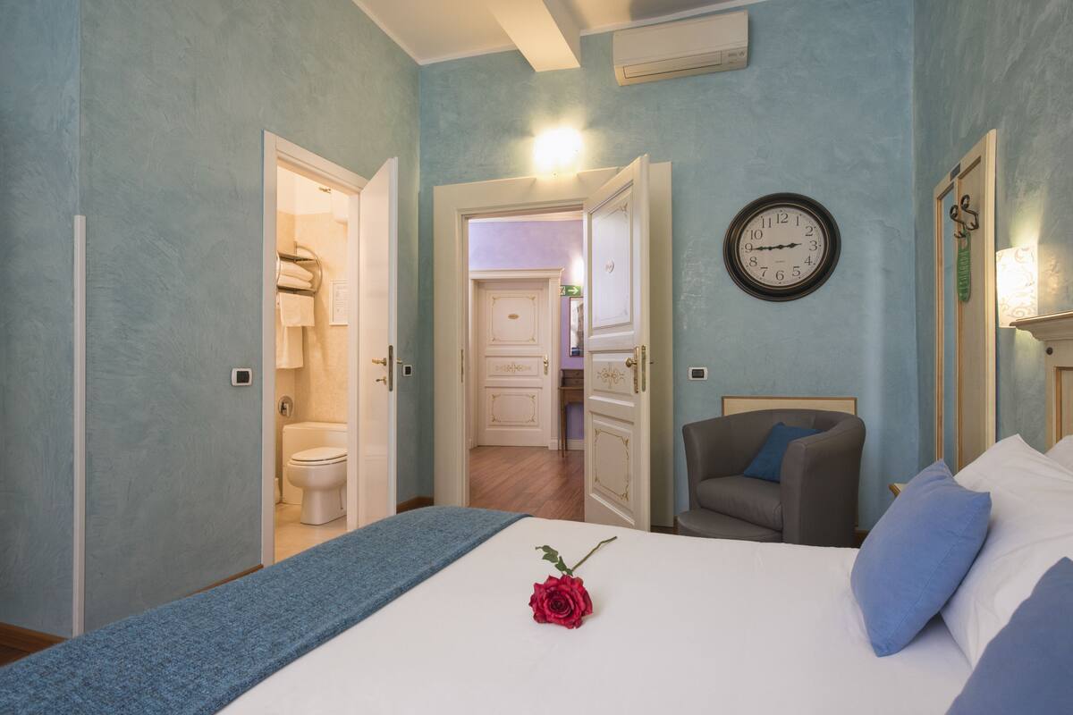 A bedroom with a bed, a chair, a clock, and an open door leading to a bathroom.