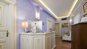 A reception area with a white counter, a desk with a computer, a lamp, and a painting on the wall. - Navona Tower Relais (Rome)