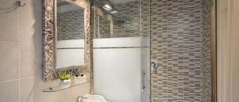 A bathroom with a glass shower enclosure, a white sink with a mirror, and a decorative plant.