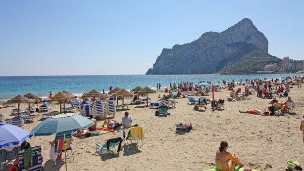 A crowded beach with numerous sun loungers and umbrellas, a prominent rocky hill in the background, and a clear blue sky. - Villas Costa Calpe - Andrómeda (Benissa)