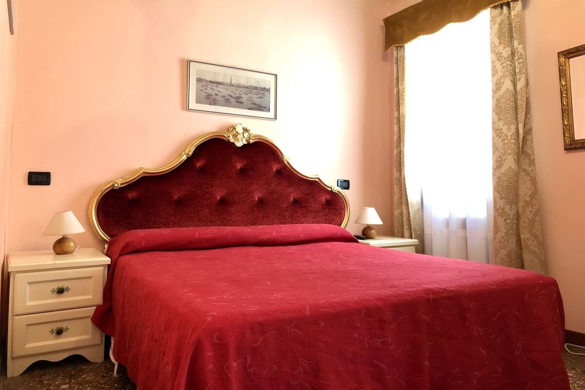 A bedroom with a large bed, two bedside tables, a nightstand, a framed picture on the wall, and a window with curtains.