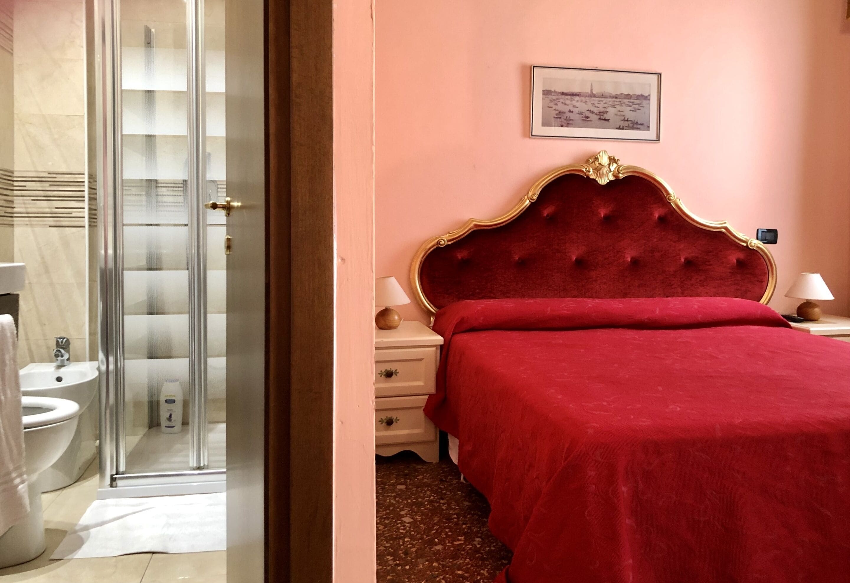 Une chambre d’hôtel dotée d’une douche en verre, d’un lit recouvert d’un tissu rouge, d’une table de chevet avec des lampes et d’un tableau encadré au mur.