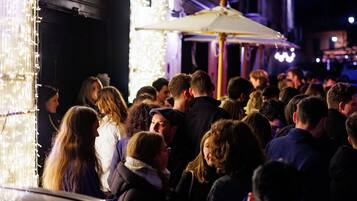 Un espace extérieur animé, où une foule de personnes s’est rassemblée autour d’un bâtiment, peut-être un restaurant ou un lieu d’événements, agrémenté de guirlandes lumineuses et de parasols.