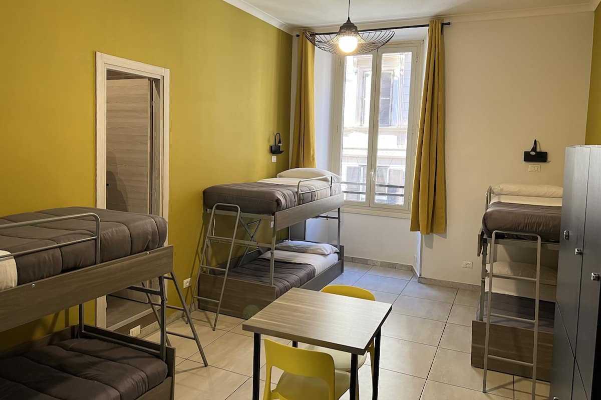 A dormitory room with bunk beds, a table, chairs, a hanging light fixture, and a window with curtains.