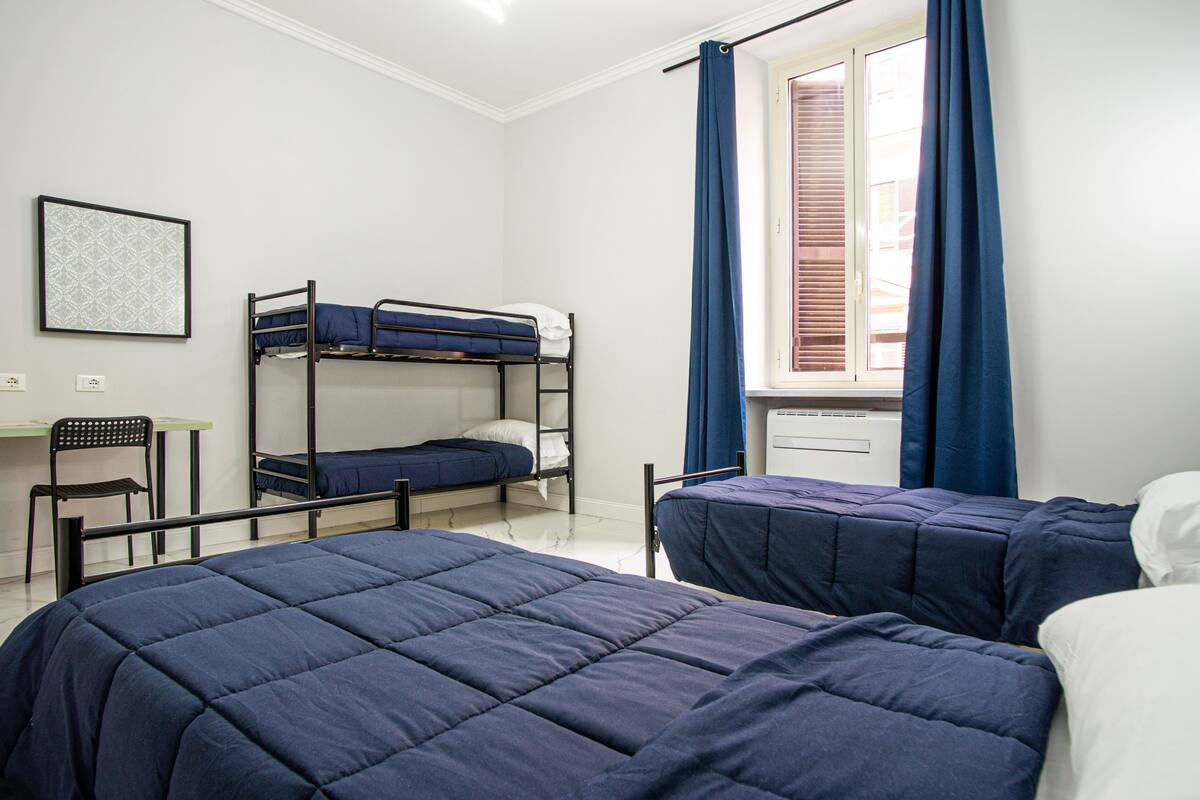 A dormitory room with bunk beds, a desk, and a window with curtains.