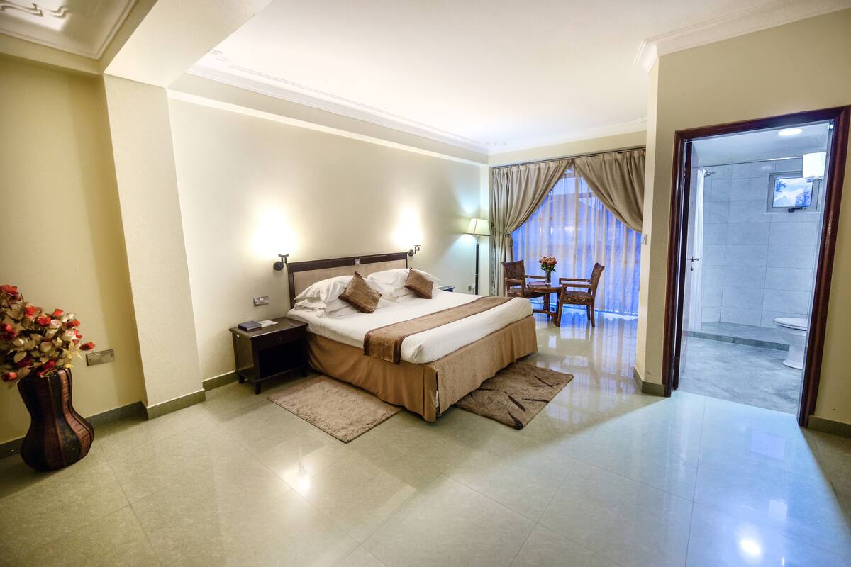 Ein Hotelzimmer mit einem großen Bett, einem Nachttisch, einem Sessel, einem kleinen Tisch mit einer Vase und Blick ins Badezimmer.