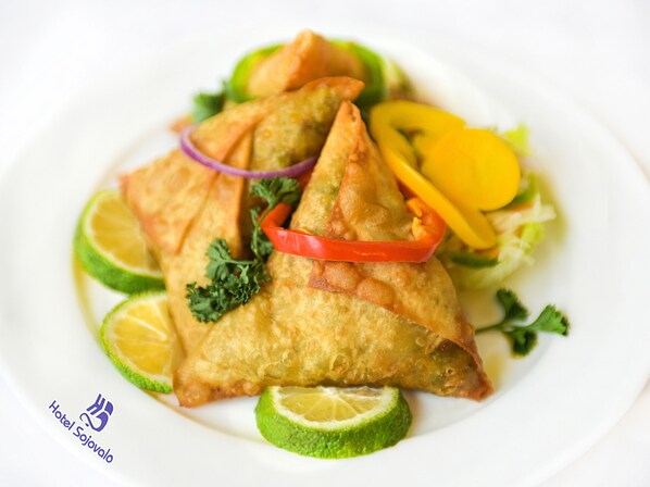 A plate of samosas garnished with lime slices, red onion, and bell pepper, with a logo for Hotel Sojovalo.