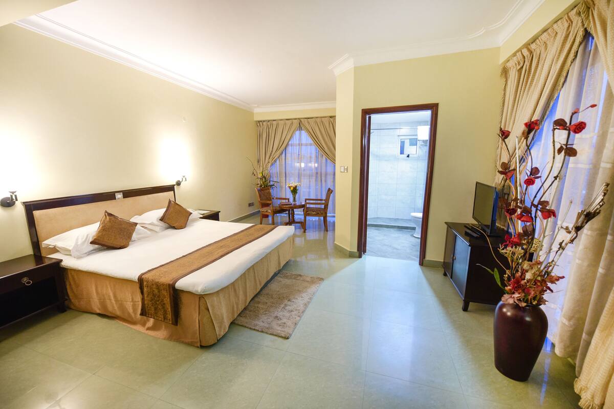 Ein Hotelzimmer mit einem großen Bett, einem Schreibtisch, einem Sessel, einem Fernseher und einer Vase mit Blumen.