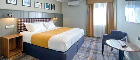 A hotel room with a large bed, a bedside table, a chair, a small table, and a window with curtains.
