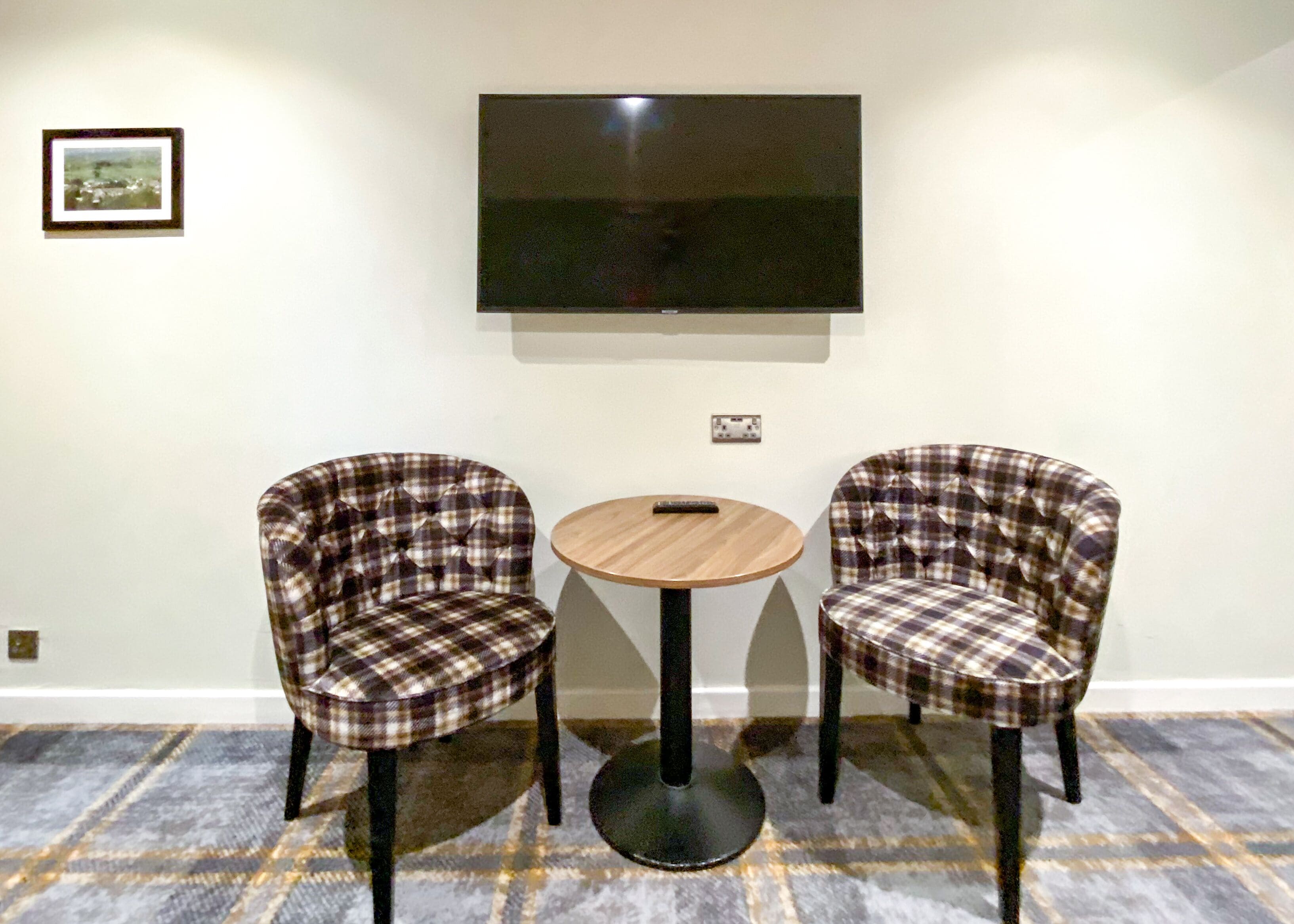 Una habitación con dos sillones a cuadros, una mesita redonda pequeña, un televisor de pantalla plana en la pared y piso alfombrado.