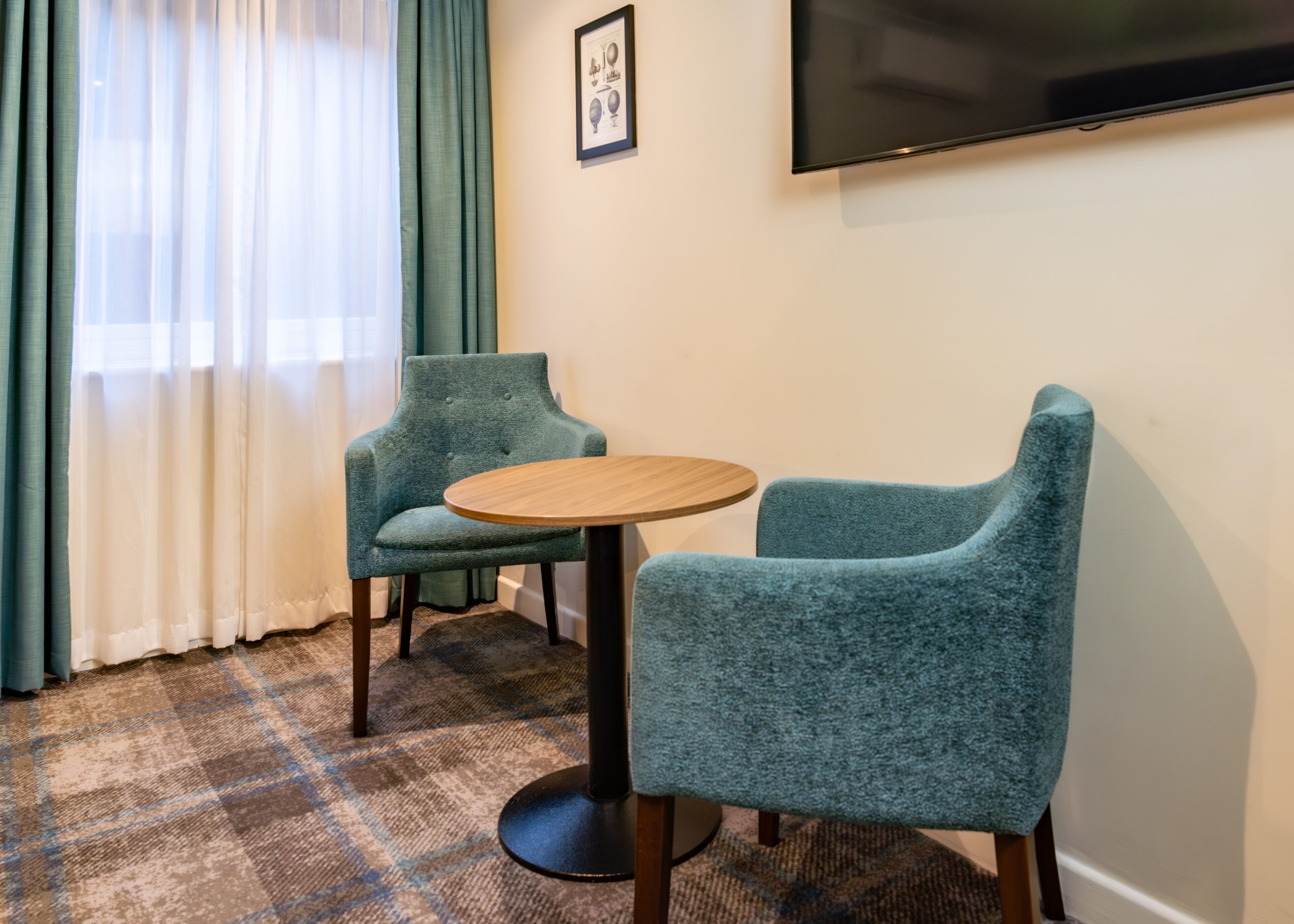 A room with two teal armchairs, a round wooden table, a large flat-screen TV mounted on the wall, and a window with sheer white curtains.
